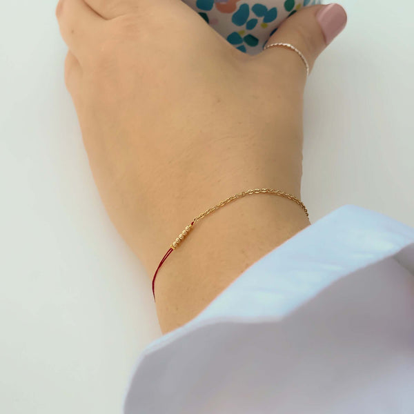 Close-up of hand wearing delicate bracelet of gold chain and red thread with gold beads detail at center.