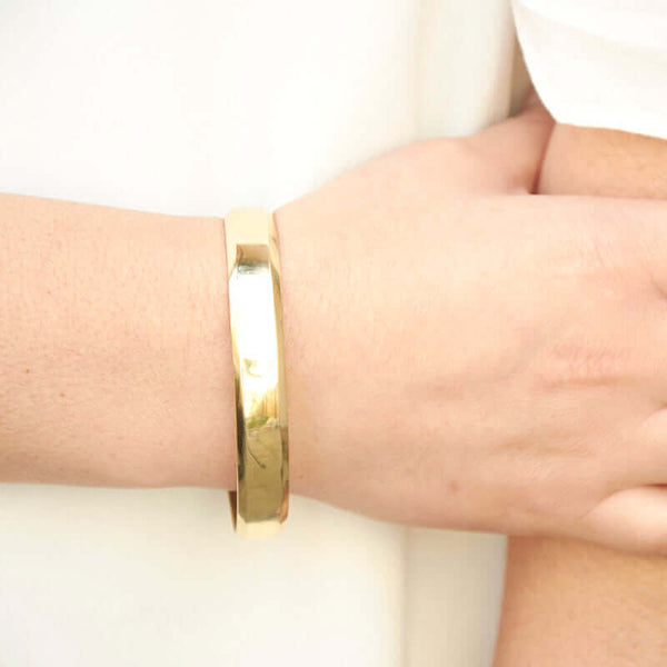 Close-up of hand wearing heavy cuff bracelet with chamfered edges and overlapping ends, in polished brass.