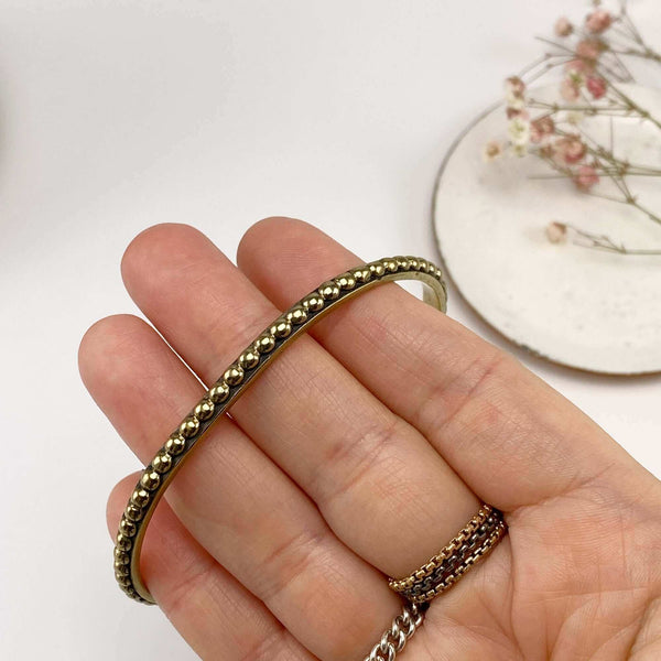 Close-up of fingers holding a polished brass beaded bangle bracelet.