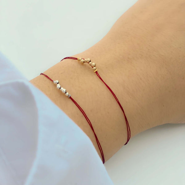 Close-up of hand wearing 2 delicate red thread bracelets, one with gold and one with silver beads detail at center.