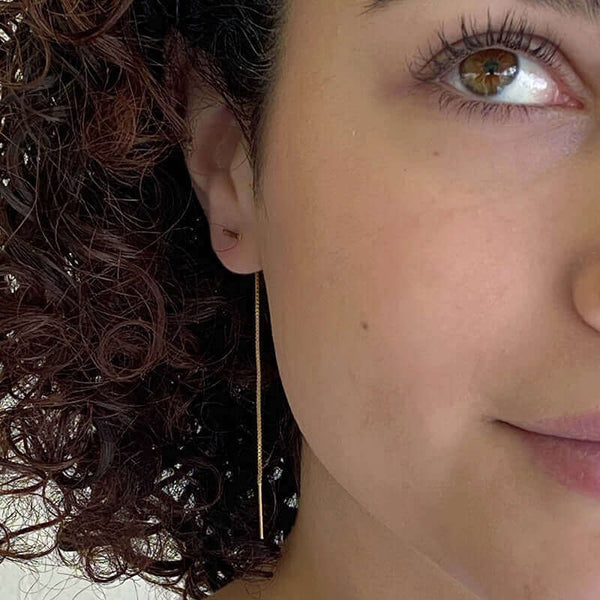 Close up front view of woman wearing gold threader earrings, small bar with fine gold chain ending in bar.