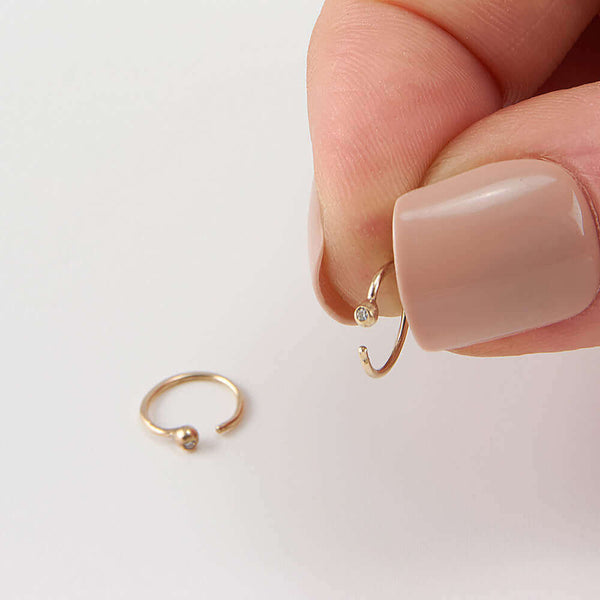 Fingers holding on of a pair of small gold hoop earring, tiny ball detail in front with inset diamond.