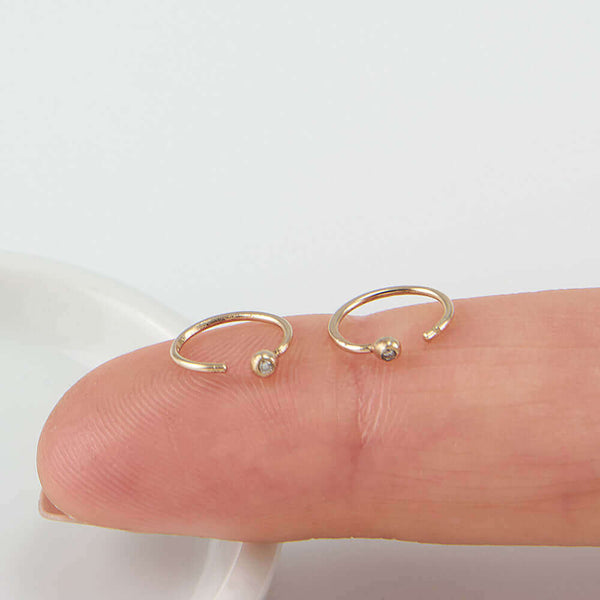 Pair of small gold hoop earring, tiny ball detail in front with inset diamond, resting on a finger.