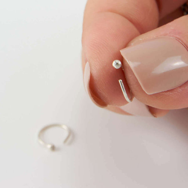 Fingers holding on of a pair of small silver hoop earring, tiny ball detail in front with inset diamond.