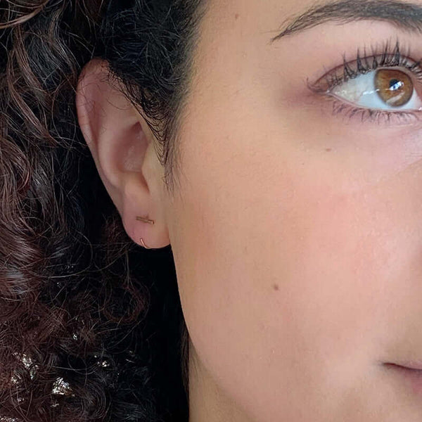 Close up front view of woman wearing gold earrings, flat bar in front with curved wire hoop.