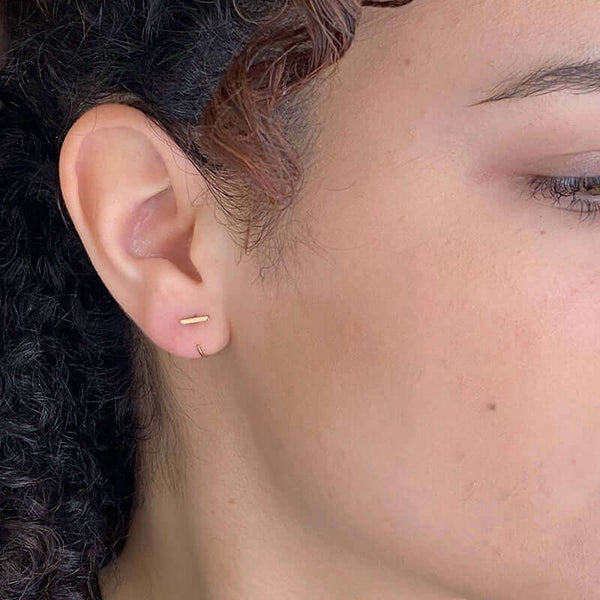 Close up side view of woman wearing gold earrings, flat bar in front with curved wire hoop.