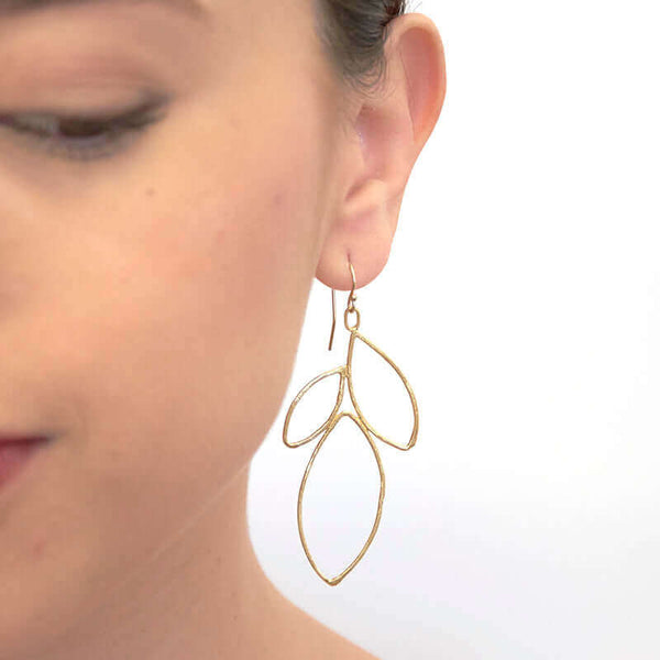 Close up front view of woman wearing gold earrings shaped like outline of group of leaves.
