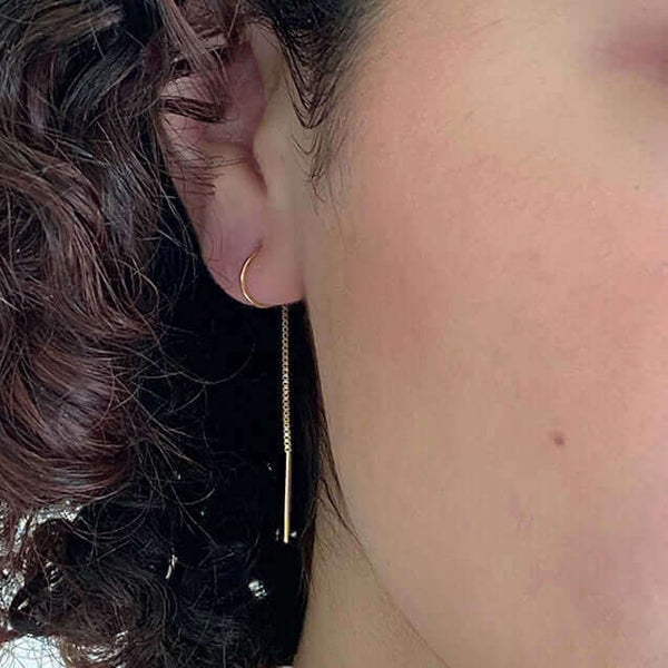 Close up front view of woman wearing gold earrings, with open hoop with thin chain hanging down.