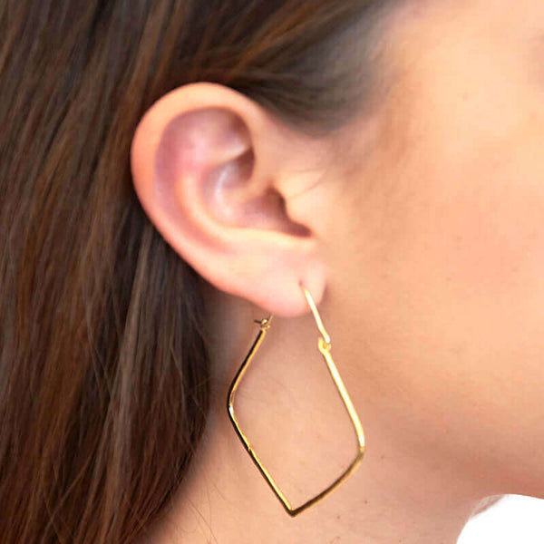 Close up side view of woman wearing pair of gold geometric rhombus shaped hoop earrings.