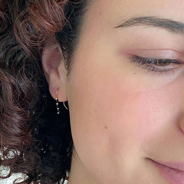 Close up front view of woman wearing pair of small gold wire earrings with black and gold beads.