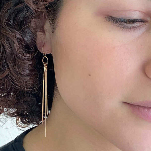 Close up front view of woman wearing pair of gold dangle earrings with three varied length square wires.