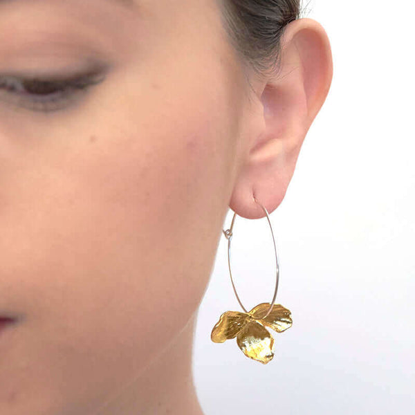 Close up front view of woman wearing pair of earrings of gold orchid petals on a silver hoop.