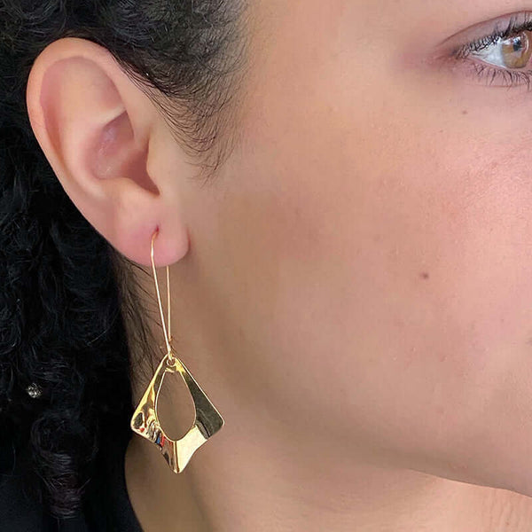 Close up side view of woman wearing pair of gold earrings, flat diamond shape with oval cutout on french earwire.