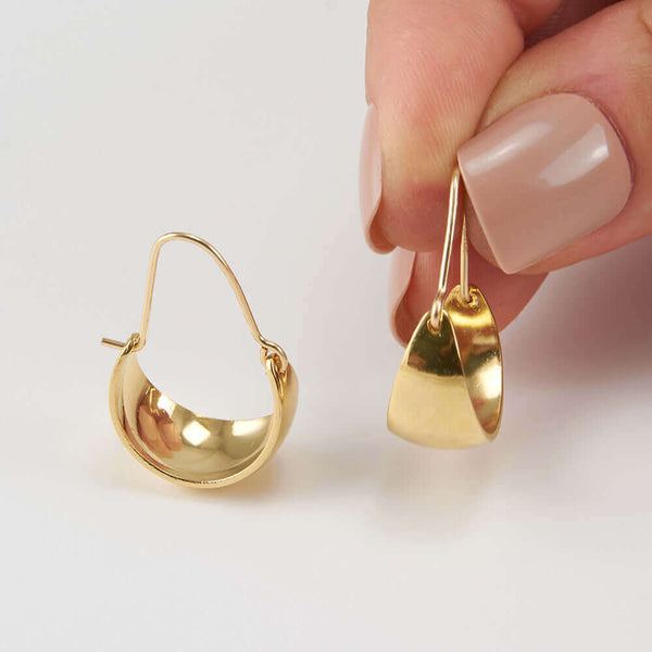 Fingers holding one of a pair of gold convex curved hoop earrings.