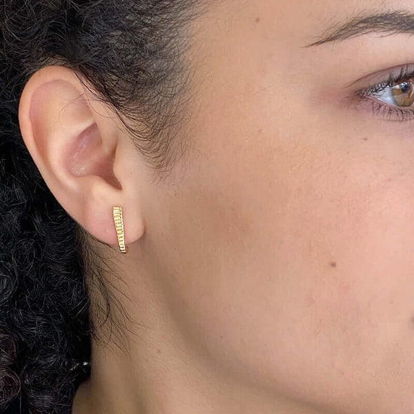 Close-up side view of woman wearing a pair of gold earrings, curved spike with serrations on posts that hooks around earlobe.