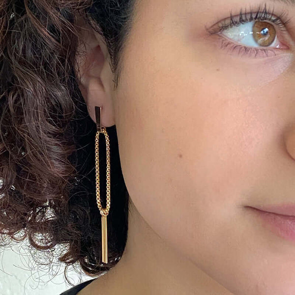Close-up front view of woman wearing a pair of gold earrings, chain hoop with 3 hanging bars on posts.