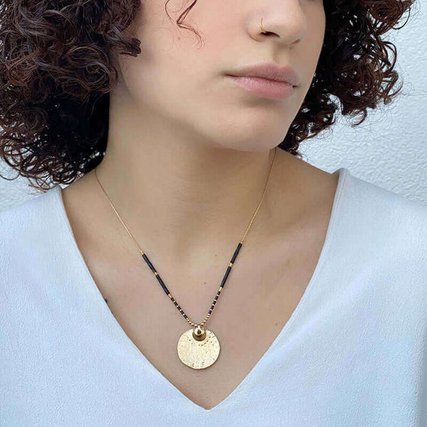 Woman wearing gold necklace with round medallion and black beads on either side, hanging short.