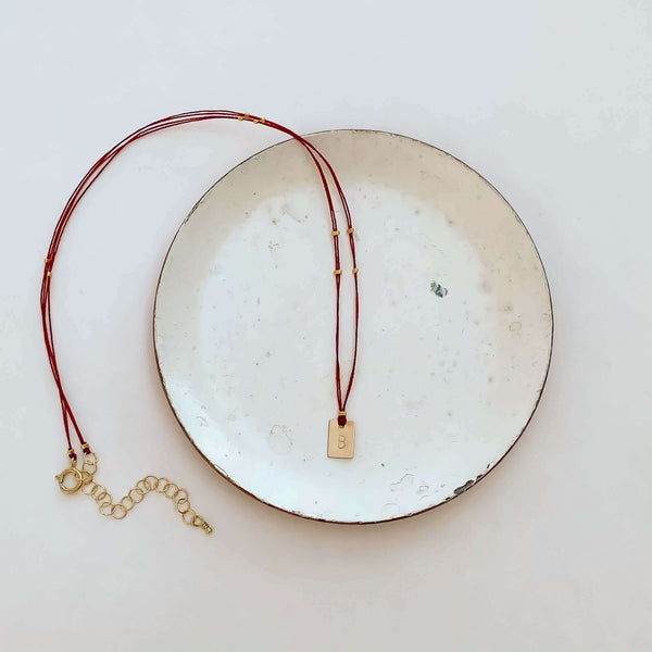 Delicate red thread necklace with gold accents and square pendant on small plate.