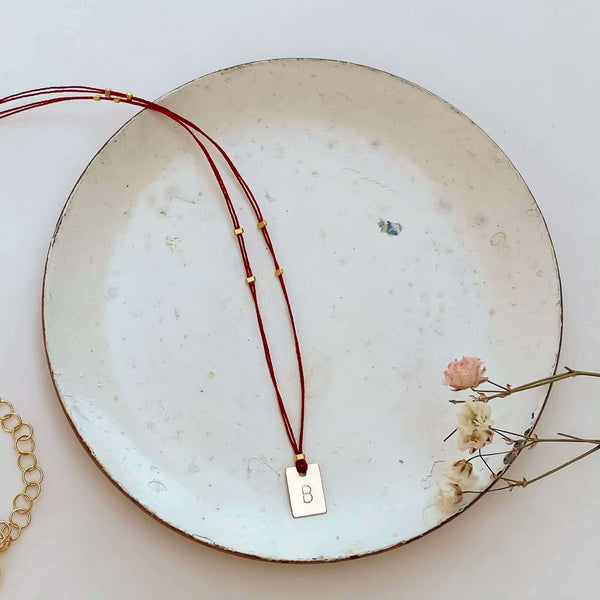Delicate red thread necklace with gold accents and square pendant on small plate.