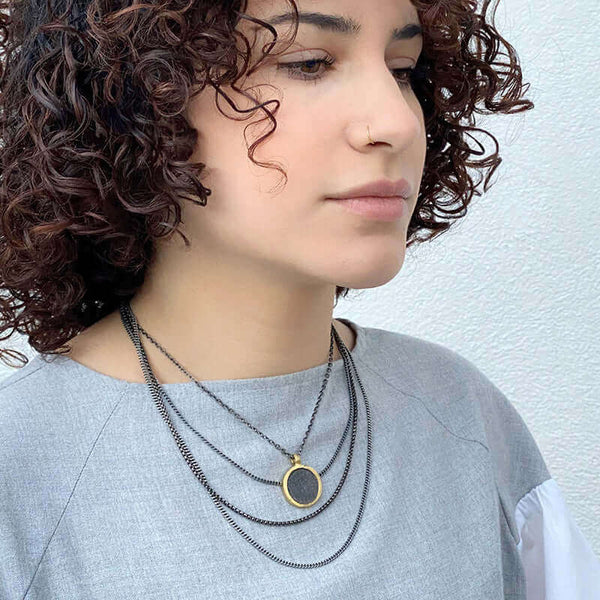 Woman wearing dark multi-chain necklace with round coin shaped etched brass pendant, worn doubled.