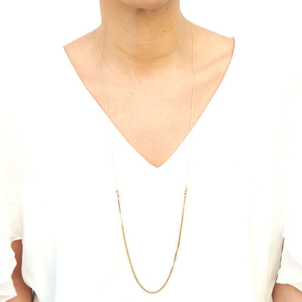Woman wearing delicate gold chain necklace with box chain front, and slider for clasp, worn long.