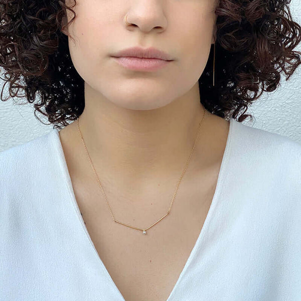 Woman wearing a delicate gold chain necklace, wide v pendant with white pearl in middle.