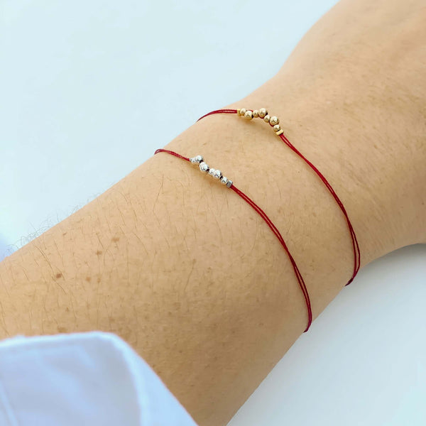 Close-up of hand wearing 2 delicate red thread bracelets, one with gold and one with silver beads detail at center.