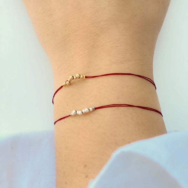 Close-up of hand wearing 2 delicate red thread bracelets, one with gold and one with silver beads detail at center.