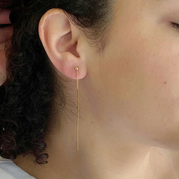 Close-up side view of woman wearing gold dangle earrings, chain with thin bar on ear post.