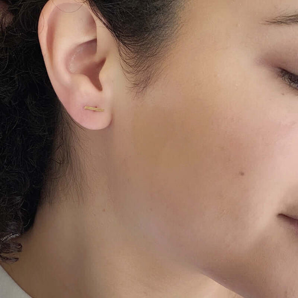 Close-up side view of woman wearing gold bar earrings on earpost horizontally.
