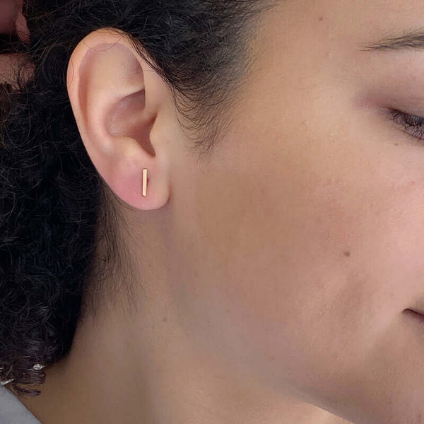 Close-up side view of woman wearing gold bar earrings on earpost vertically.