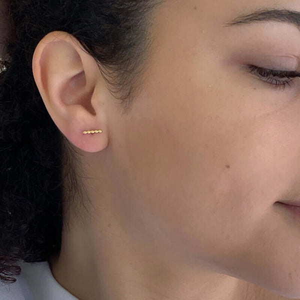 Close-up side view of woman wearing gold beaded bar earrings on earpost.