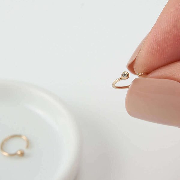 Fingers holding on of a pair of small gold hoop earring, tiny ball detail in front with inset diamond.