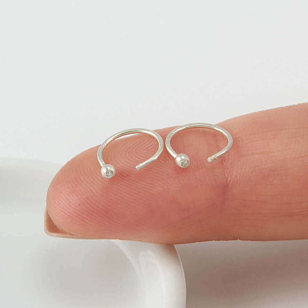 Pair of small silver hoop earring, tiny ball detail in front with inset diamond, resting on a finger.