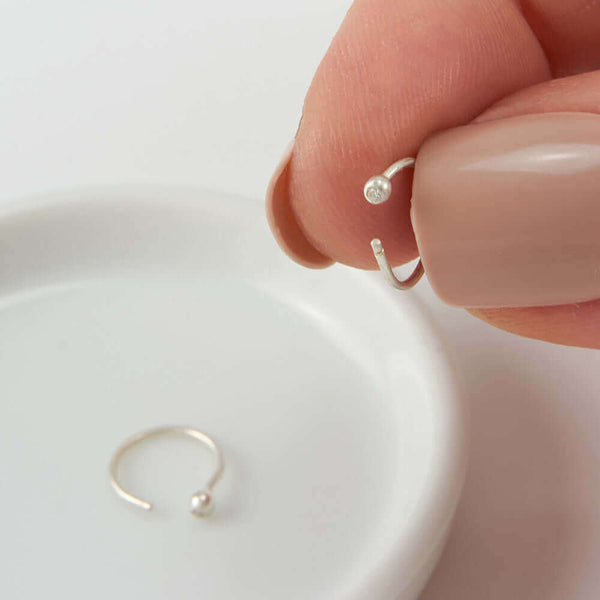 Fingers holding on of a pair of small silver hoop earring, tiny ball detail in front with inset diamond.