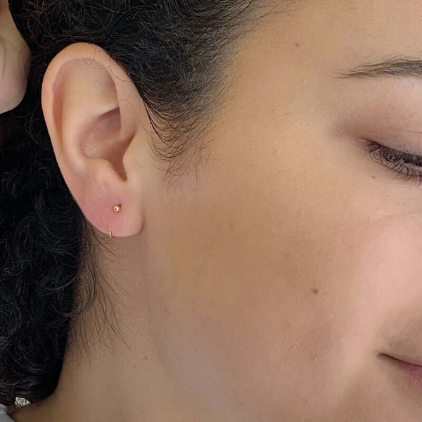 Close up side view of woman wearing small gold hoop earring with a tiny ball detail in front, facing in.