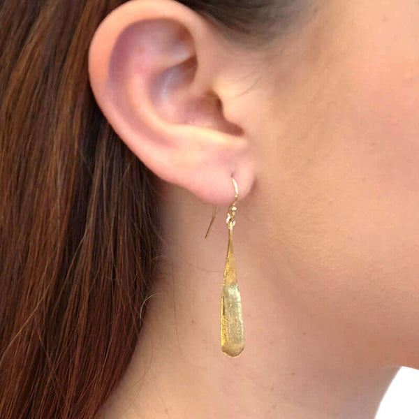 Close up front view of woman wearing gold earrings shaped like elongated teardrop.