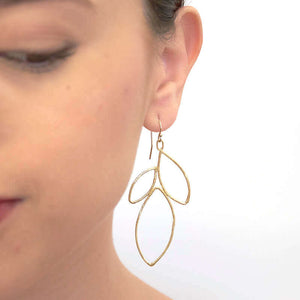 Close up front view of woman wearing gold earrings shaped like outline of group of leaves.