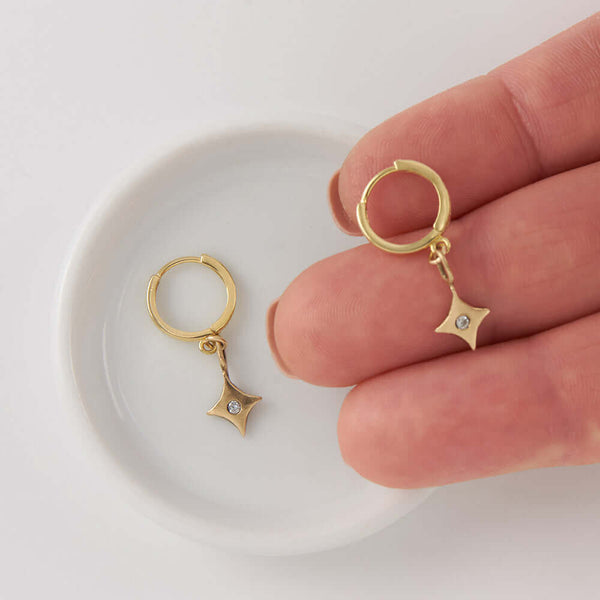 Fingers holding one of a pair of 4 point gold star earrings on hoop clasp.