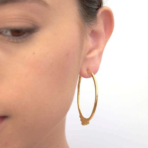 Close up front view of woman wearing pair of gold geometric native motif hoop earrings.