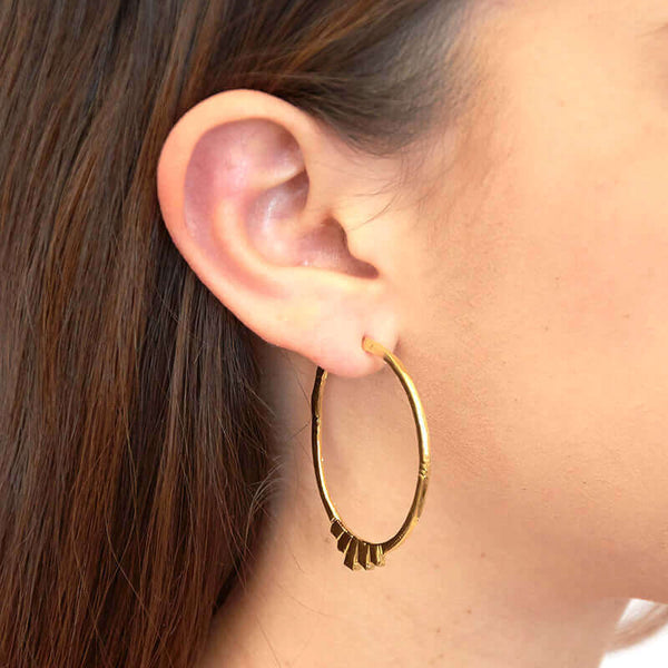 Close up side view of woman wearing pair of gold geometric native motif hoop earrings.