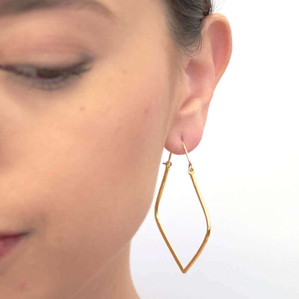 Close up front view of woman wearing pair of gold geometric rhombus shaped hoop earrings.