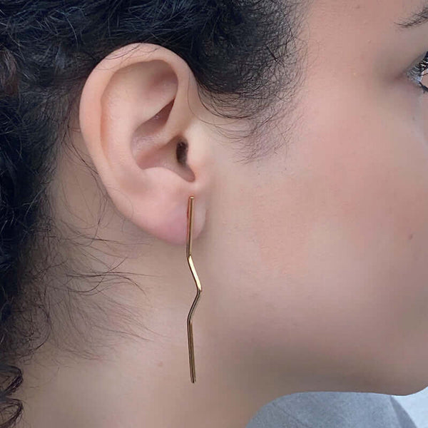 Close-up side view of woman wearing pair of gold wire earrings with geometric zig zag design.