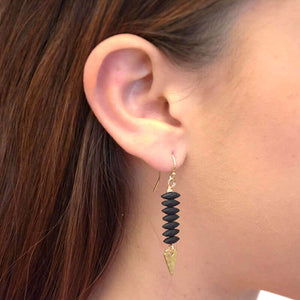 Close up side view of woman wearing pair of gold earrings with black beads and hammered arrowhead detail at end.