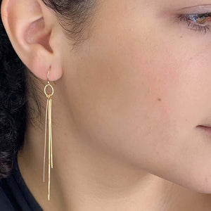 Close up side view of woman wearing pair of gold dangle earrings with three varied length square wires.
