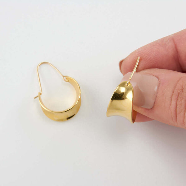 Fingers holding one of a pair of gold concave curved hoop earrings.