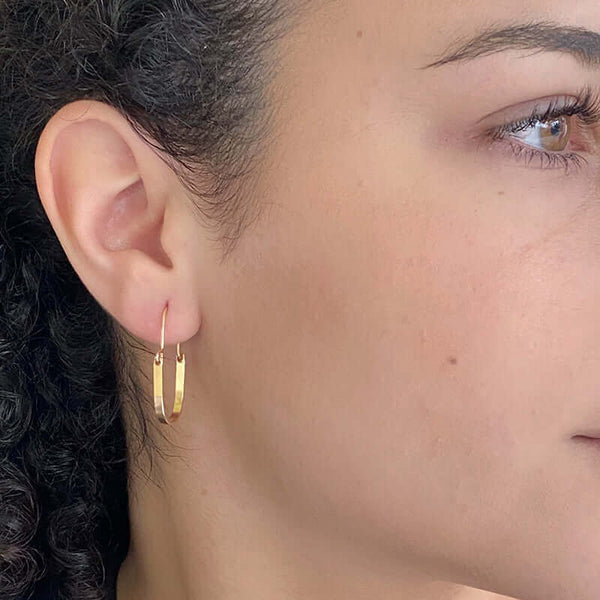 Close up side view of woman wearing pair of gold hoop style earrings with curved bar on wire.