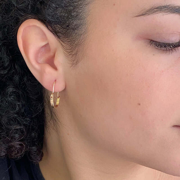 Close up side view of woman wearing pair of small gold hoop style earrings with curved bar on wire.