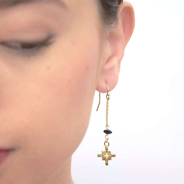 Close up front view of woman wearing pair of gold flat chain earrings with geometric cross at end.