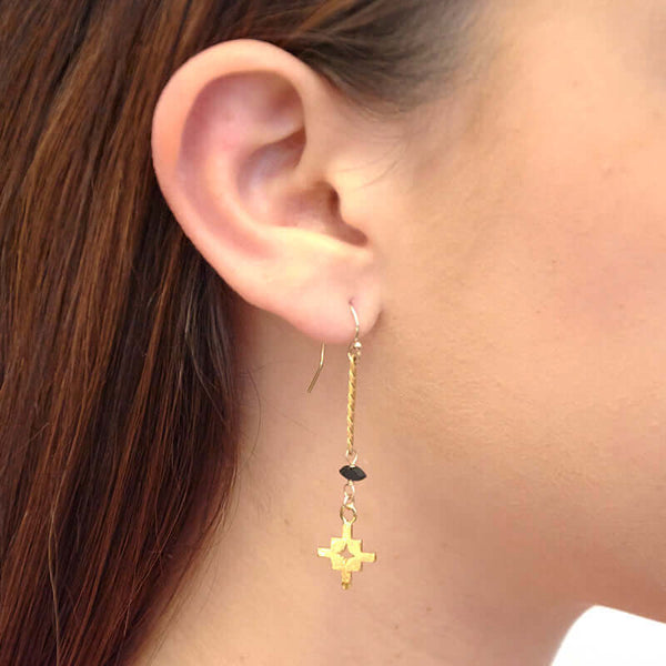 Close up side view of woman wearing pair of gold flat chain earrings with geometric cross at end.